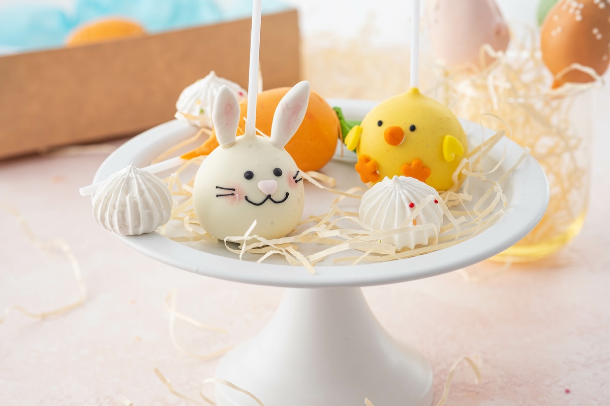 Cake pop de conejito de Pascua y cake pop de pollito en un plato de tarta con más decoración.