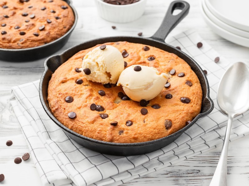 Una skillet cookie, es decir, una galleta en sartén, con chispas de chocolate en una sartén, decorada con dos bolas de helado. Junto a ella, una cuchara y otra sartén con una galleta.