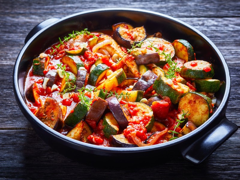 Un guiso de verduras con berenjenas, calabacín, tomates, ajo y cebollas, servido en un tazón de cerámica negro y adornado con hierbas. El tazón está sobre una superficie de madera oscura.