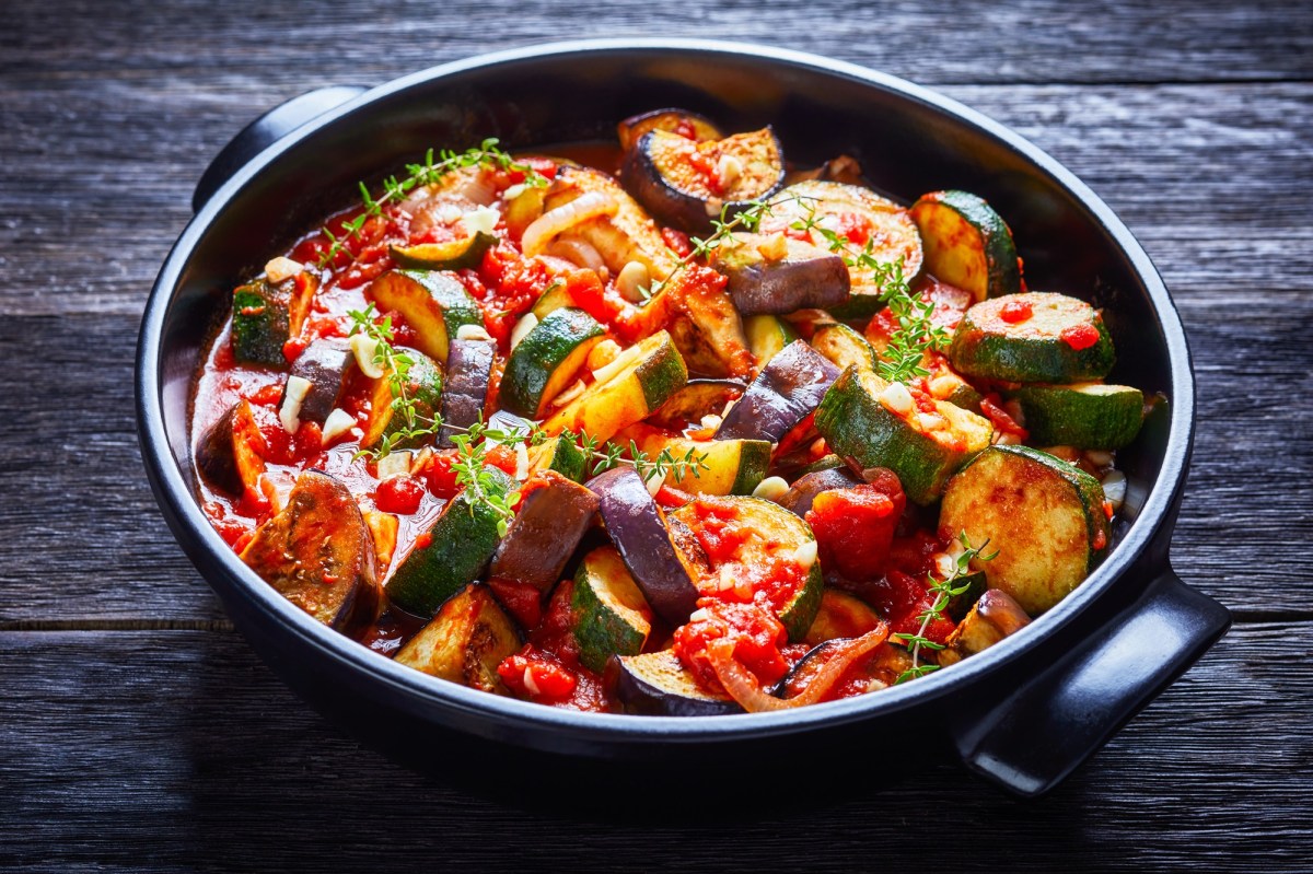 Un guiso de verduras con berenjenas, calabacín, tomates, ajo y cebollas, servido en un tazón de cerámica negro y adornado con hierbas. El tazón está sobre una superficie de madera oscura.