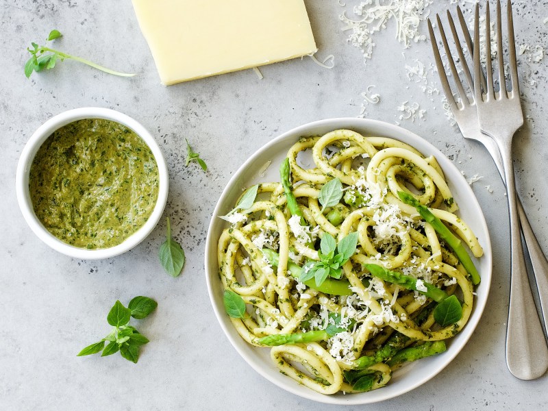 Un plato de espaguetis con pesto de espárragos, al lado un tazón de pesto. En la parte superior del borde de la imagen hay un trozo de parmesano.