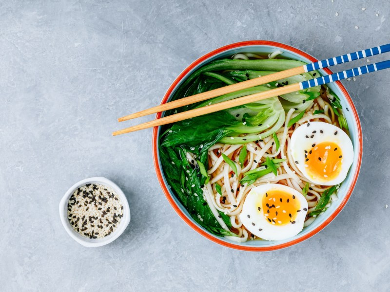 Un tazón de ramen de mantequilla de cacahuate con medio huevo y pak choi en vista superior, con palillos encima y un tazón de sésamo al lado.