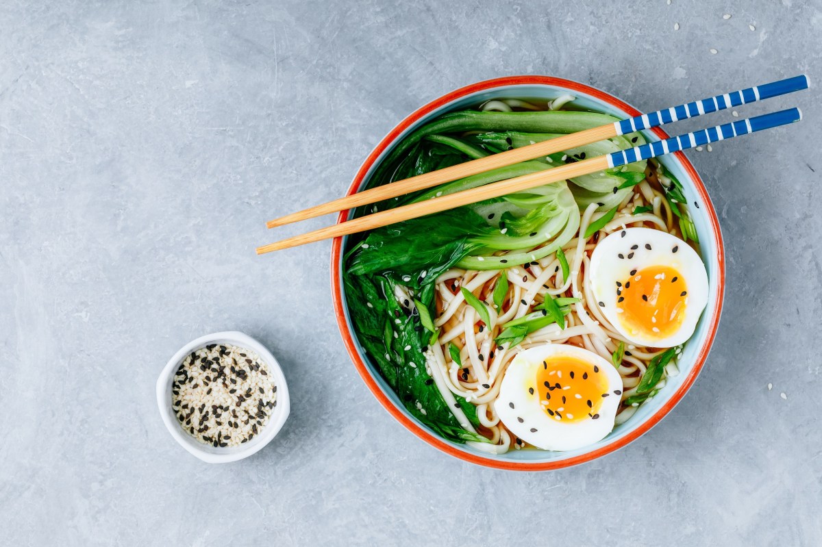 Un tazón de ramen de mantequilla de cacahuate con medio huevo y pak choi en vista superior, con palillos encima y un tazón de sésamo al lado.