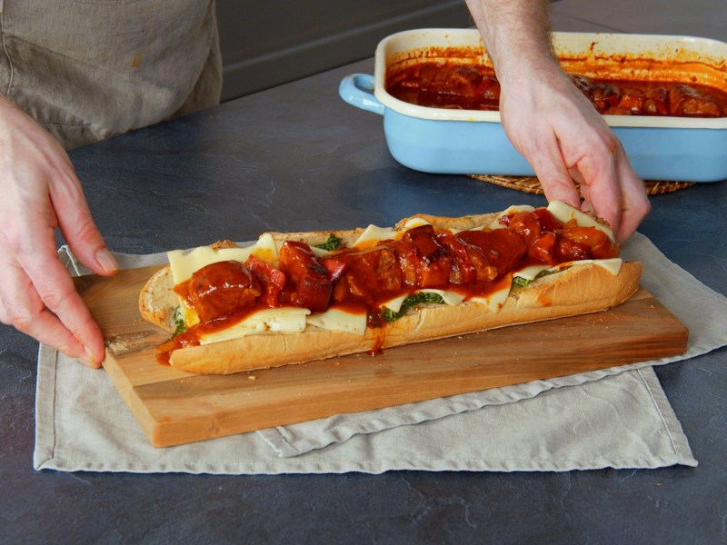 Un sándwich de pinchos morunos sobre una tabla de madera. Debajo de la tabla hay manteles individuales de tela. Junto al sándwich de pinchos morunos hay una fuente de horno azul claro con salsa de pinchos morunos y más pinchos morunos. Se pueden ver dos manos cogiendo el sándwich.