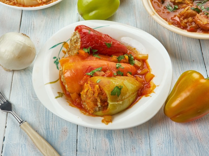 3 pimientos rellenos húngaros con salsa de tomate en un plato, junto a un tenedor, un pimiento y una cebolla, así como una olla con salsa de tomate.