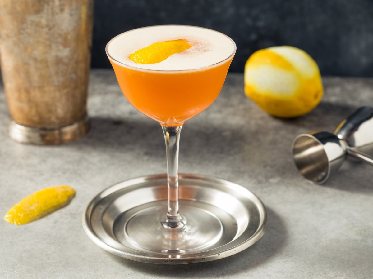 Earl Grey Sour en una copa de cóctel sobre una bandeja de plata. Está decorado con una cáscara de limón, detrás hay un limón.
