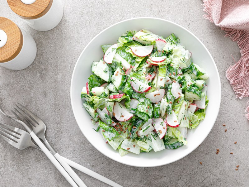 Tazón de ensalada con rábanos y pepinos sobre una mesa clara y cubiertos.