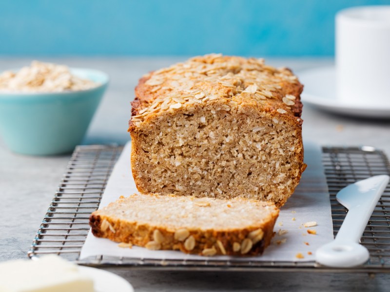 Un pan de avena cortado en una tabla, junto a él un cuchillo blanco, al fondo desenfocado un tazón con hojuelas de avena.