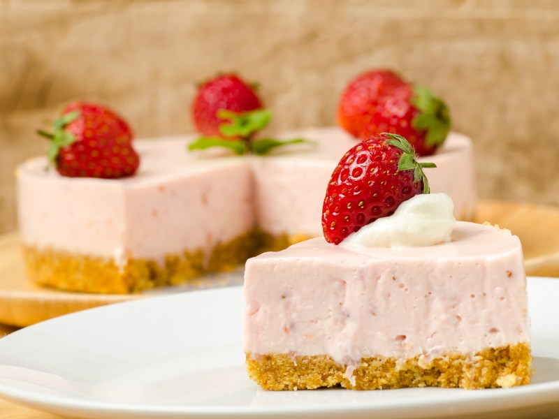 Un trozo de pastel de mousse de fresa, decorado con una fresa, en un plato, con el pastel cortado al fondo.