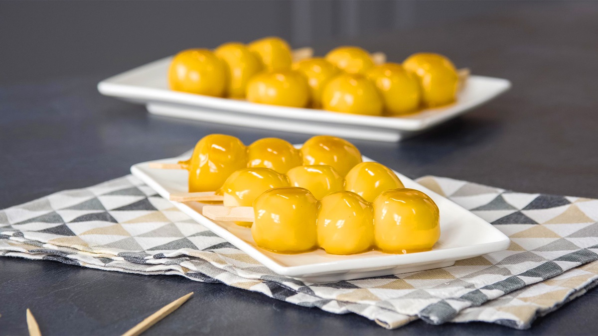 Tres brochetas de dango se encuentran en un plato blanco. Al fondo se ven más brochetas de dango. Están cubiertas con un glaseado dorado.