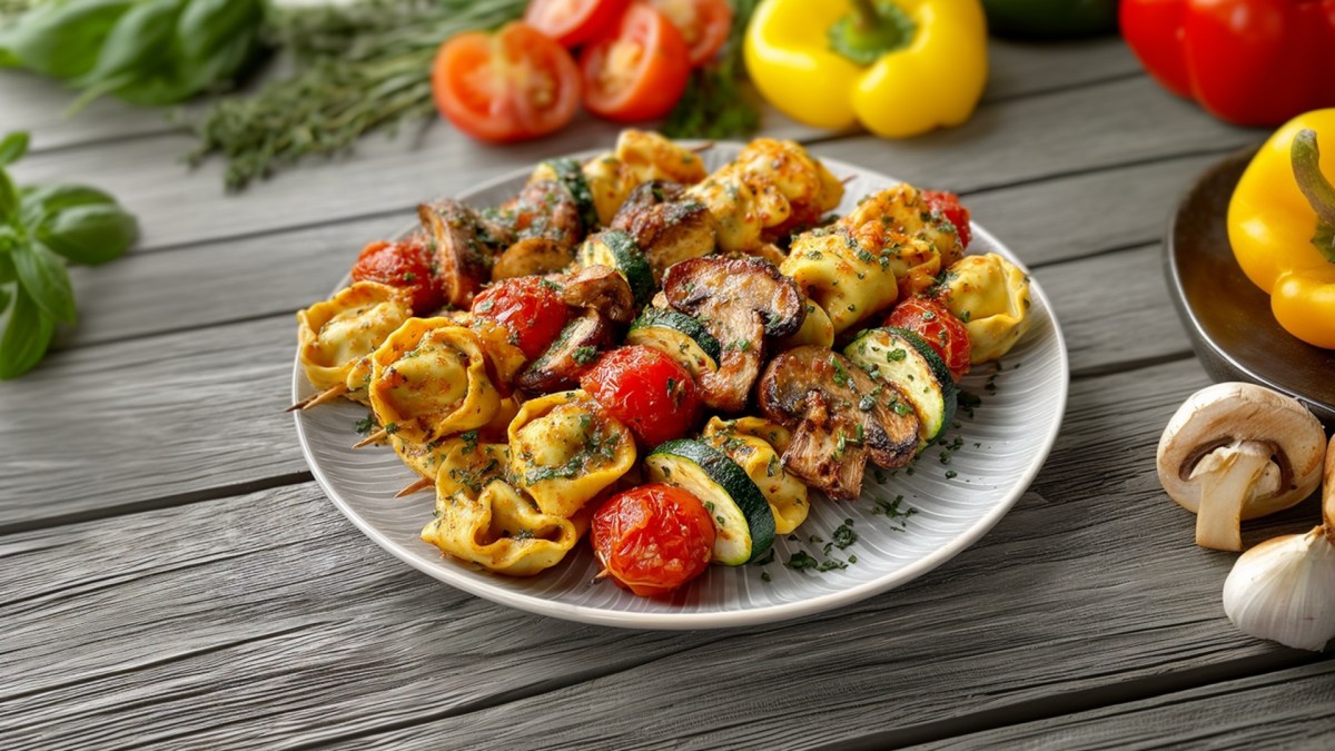 Un plato con brochetas de tortelloni a la parrilla. El plato está sobre una base de madera. Alrededor hay pimientos amarillos, tomates y hierbas frescas esparcidas.