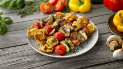 Un plato con brochetas de tortelloni a la parrilla. El plato está sobre una base de madera. Alrededor hay pimientos amarillos, tomates y hierbas frescas esparcidas.