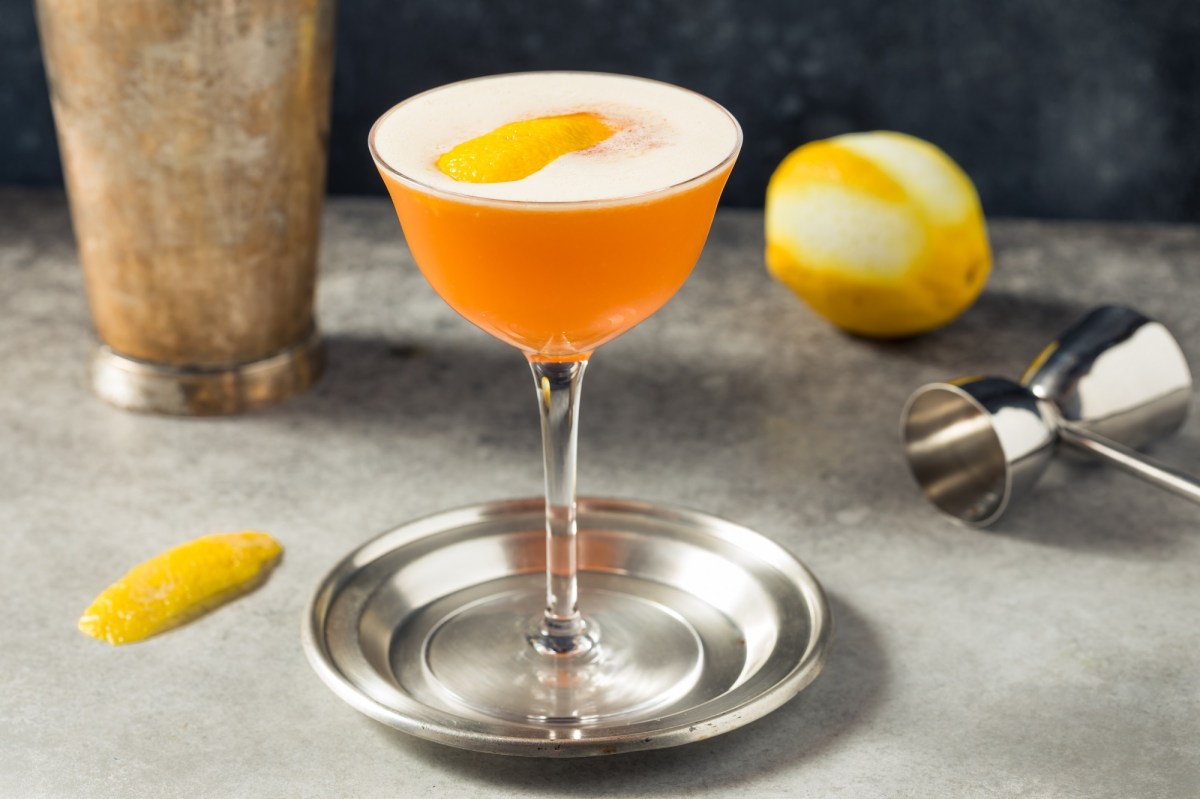 Earl Grey Sour en una copa de cóctel sobre una bandeja de plata. Está decorado con una cáscara de limón, detrás hay un limón.