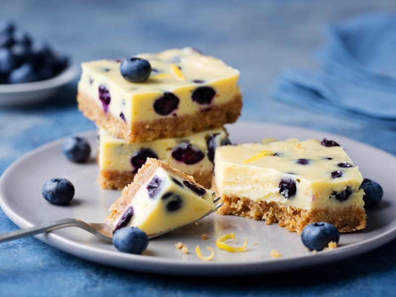 Varias porciones de pastel de queso con limón y arándanos sin azúcar en un plato. Alrededor hay arándanos.