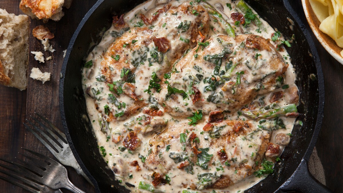 Pollo en salsa de parmesano en una sartén, fotografiado desde arriba. Junto a él hay cubiertos y trozos de pan.